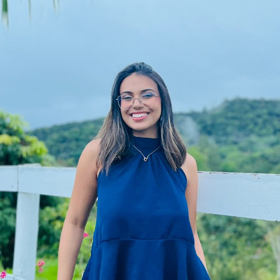 Natália Paixão, sorrindo, encostada numa cerca branca com um morro verde ao fundo